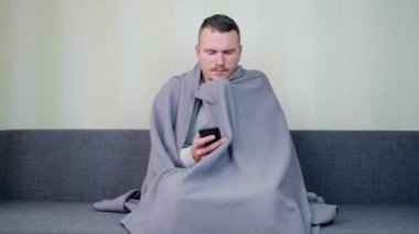 Hasta adam battaniyeyle siper alıyor ve akıllı telefon kullanıyor. Üzgün genç adam kanepede oturuyor ve ateşler içinde titriyor. Akut Solunum Virüsü Enfeksiyonu. Soğuk algınlığı. 4K 'da kendi kendini tedavi ediyor.