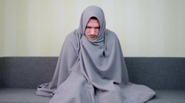 Hasta adam ateşten titriyor ve battaniyeyle korunuyor. Üzgün genç adam yüksek ateşle kanepede oturuyor. Akut Solunum Virüsü Enfeksiyonu. Soğuk algınlığı. 4k görüntü.