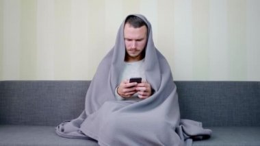 Battaniyeyle kaplanmış hasta adam akıllı telefon kullanıyor. Üzgün genç adam yüksek ateşle kanepede oturuyor. Akut Solunum Virüsü Enfeksiyonu. Soğuk algınlığı. Evde kendi kendini tedavi ediyor. 4k görüntü