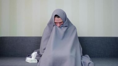 Battaniyeyle kaplanmış hasta bir adam. Üzgün genç adam yüksek ateşle kanepede oturuyor. Akut Solunum Virüsü Enfeksiyonu. Soğuk algınlığı. Evde kendi kendini tedavi ediyor. 4k görüntü.