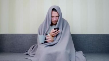 Battaniyeyle kaplanmış hasta bir adam bir fincan sıcak çay içer ve akıllı telefon kullanır. Üzgün genç adam yüksek ateşle kanepede oturuyor. Akut Solunum Virüsü Enfeksiyonu. Soğuk algınlığı. 4k görüntü.