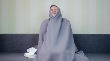 Battaniyeyle kaplanmış hasta bir adam peçeteyi alır ve içine hapşırır. Üzgün genç adam yüksek ateşle kanepede oturuyor. Akut Solunum Virüsü Enfeksiyonu. Soğuk algınlığı. 4k görüntü.