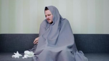 Battaniyeyle kaplanmış hasta bir adam peçeteyi alır ve içine hapşırır. Üzgün genç adam yüksek ateşle kanepede oturuyor. Akut Solunum Virüsü Enfeksiyonu. Soğuk algınlığı. 4k görüntü.