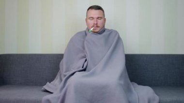Battaniyeyle kaplanmış hasta adam sıcaklığı ölçer. Ateşli genç adam kanepede oturuyor. Akut Solunum Virüsü Enfeksiyonu. Soğuk algınlığı. Evde kendi kendini tedavi ediyor. 4k görüntü.