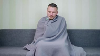 Battaniyeyle kaplanmış hasta adam sıcaklığı ölçer ve sonuç karşısında şaşırır. Genç adamın ateşi çıktı. Akut Solunum Virüsü Enfeksiyonu. Soğuk algınlığı. Evde kendi kendini tedavi ediyor. 4k görüntü.