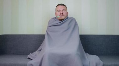 Battaniyeyle kaplanmış hasta adam sıcaklığı ölçer. Üzgün genç adam ateşler içinde titriyor. Akut Solunum Virüsü Enfeksiyonu. Soğuk algınlığı. Evde kendi kendini tedavi ediyor. 4k görüntü.