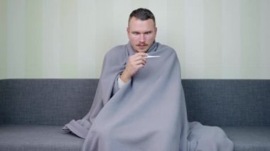 Battaniyeyle kaplanmış hasta adam sıcaklığı ölçer ve sonuç karşısında şaşırır. Genç adamın ateşi çıktı. Akut Solunum Virüsü Enfeksiyonu. Soğuk algınlığı. Evde kendi kendini tedavi ediyor. 4k görüntü.