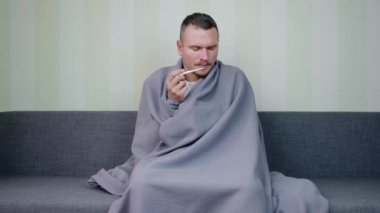 Battaniyeyle kaplanmış hasta adam sıcaklığı ölçer ve iyi bir sonuçtan zevk alır. Genç adamın ateşi çıktı. Akut Solunum Virüsü Enfeksiyonu. Soğuk algınlığı. Evde kendi kendini tedavi ediyor. 4k görüntü.