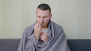 Battaniyeyle kaplanmış hasta adam burun spreyiyle tedavi edilir. Üzgün genç adam yüksek ateşle kanepede oturuyor. Akut Solunum Virüsü Enfeksiyonu. Soğuk algınlığı. 4K 'da kendi kendini tedavi ediyor.