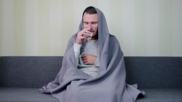 毛布で覆われた病人は丸薬を飲み 水を一杯飲む 動揺した若い男がソファに座って薬を飲んでいる 急性呼吸器ウイルス感染症 風邪のインフルエンザ自己投薬4K — ストック動画