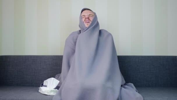 毛布で覆われた病気の男はナプキンを取り それにくしゃみをします 動揺した若い男が高温でソファに座っている 急性呼吸器ウイルス感染症 風邪のインフルエンザ4K映像 — ストック動画