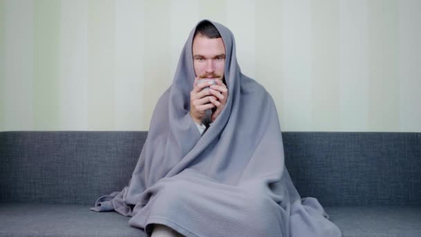 病気の男毛布で覆われてお茶のホットカップを飲む 動揺した若い男が高温でソファに座っている 急性呼吸器ウイルス感染症 風邪のインフルエンザ自宅での自己投薬 — ストック動画