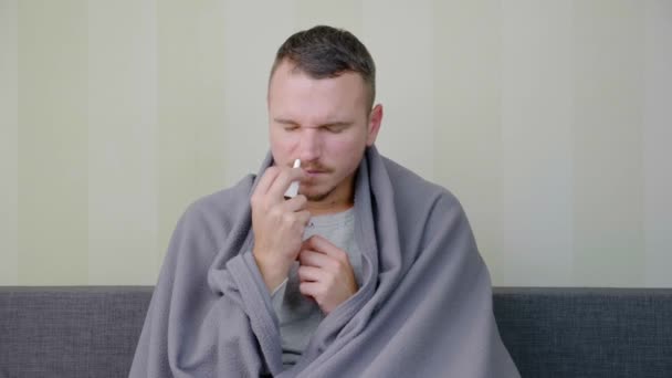 毛布で覆われた病人は鼻スプレーで治療される 動揺した若い男が高温でソファに座っている 急性呼吸器ウイルス感染症 風邪のインフルエンザ自宅での自己投薬4K — ストック動画