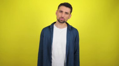 Mavi tişörtlü ve beyaz tişörtlü genç bir adam ciddi ciddi kameraya bakıyor ve başını çeviriyor. Kopyalama alanı olan sarı arkaplan. 4k yavaş çekim görüntüsü