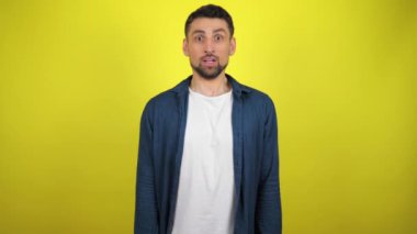 Mavi tişörtlü ve beyaz tişörtlü genç bir adam şaşırdı ve kameraya baktı. Sarı arka plan. 4k yavaş çekim görüntüsü