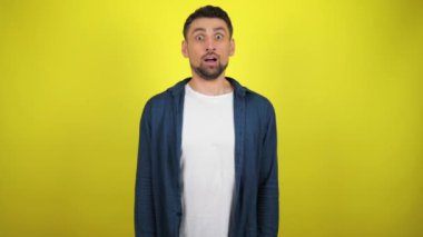Mavi tişörtlü ve beyaz tişörtlü genç bir adam başını kameraya çevirdi ve çok şaşırdı. Kopyalama alanı olan sarı arkaplan. 4k yavaş çekim görüntüsü