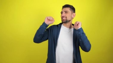 Beyaz tişörtlü ve mavi gömlekli genç bir adam sarı arka planda bir gülümsemeyle dans ediyor. 4k görüntü