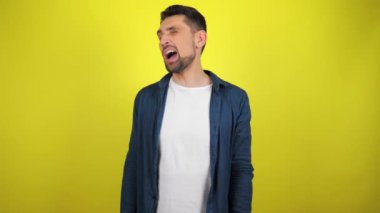 Mavi tişörtlü ve beyaz tişörtlü genç bir adam kameraya bakıyor ve göz kırpıyor. Kopyalama alanı olan sarı arkaplan. 4k yavaş çekim görüntüsü