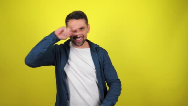白いTシャツと青いシャツを着た若い男が黄色い背景で笑顔で踊っている 4K映像 — ストック動画