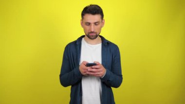 Mavi tişörtlü ve beyaz tişörtlü genç bir adam akıllı telefonu faiziyle kullanır. Kopyalama alanı olan sarı arkaplan. 4k yavaş çekim görüntüsü