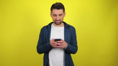 Mavi tişörtlü ve beyaz tişörtlü genç bir adam akıllı telefonu faiziyle ve gülümsemesiyle kullanır. Kopyalama alanı olan sarı arkaplan. 4k yavaş çekim görüntüsü