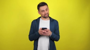 Mavi tişörtlü ve beyaz tişörtlü genç bir adam akıllı telefonu faiziyle ve gülümsemesiyle kullanır. Kopyalama alanı olan sarı arkaplan. 4k yavaş çekim görüntüsü