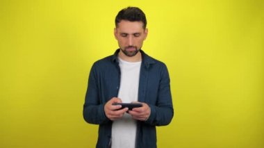 Mavi tişörtlü ve beyaz tişörtlü genç bir adam akıllı telefonda oyun oynuyor ve kaybediyor, üzülüyor. Kopyalama alanı olan sarı arkaplan. 4k yavaş çekim görüntüsü