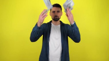 Mavi tişörtlü ve beyaz tişörtlü genç bir adam kameraya bakıyor ve havaya bir sürü para saçıyor. Kopyalama alanı olan sarı arkaplan. 4k yavaş çekim görüntüsü