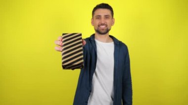 Beyaz tişörtlü genç bir adam bir hediyeyle elini kaldırıyor ve kameraya gülümsüyor, sarı arka planda. Tatil konsepti: Yeni yıl, Noel, Aziz Nicholas Günü, Sevgililer Günü, doğum günü. 4k görüntü