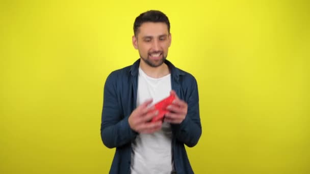 一个穿着白色T恤的年轻人手里拿着一个盒子 手里拿着礼物 看着相机 微笑着 黄色背景 假期概念 圣诞节 情人节 4K镜头 — 图库视频影像
