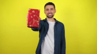 Beyaz tişörtlü genç bir adam bir hediyeyle elini kaldırıyor ve kameraya gülümsüyor, sarı arka planda. Tatil konsepti: Yeni yıl, Noel, Aziz Nicholas Günü, Sevgililer Günü, doğum günü. 4k görüntü