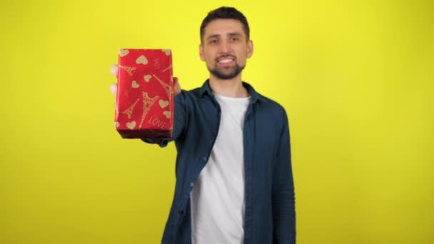 白いTシャツを着た若い男が手に贈り物を持ち カメラに向かって黄色の背景で微笑む 休日の概念 クリスマス 聖ニコラスデー バレンタインデー 誕生日 4K映像 — ストック動画