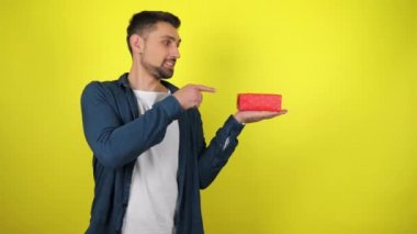 Beyaz tişörtlü genç bir adam avucunun içinde tuttuğu hediyeye parmağını gösteriyor, gülümsüyor ve sarı arka plan gibi görünüyor. Tatil konsepti: yeni yıl, Noel, doğum günü. 4kfootage