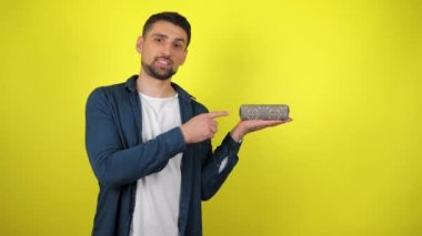 Beyaz tişörtlü genç bir adam avucunun içinde tuttuğu hediyeye parmağını gösteriyor, gülümsüyor ve sarı arka plan gibi görünüyor. Tatil konsepti: yeni yıl, Noel, doğum günü. 4kfootage