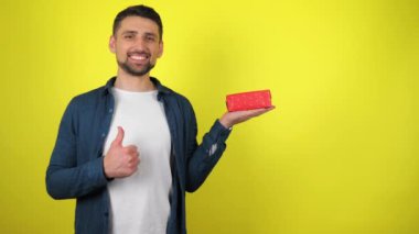 Beyaz tişörtlü genç bir adam avucunda bir hediye tutuyor, gülümsüyor ve sarı arka plan gibi görünüyor. Tatil konsepti: Yeni yıl, Noel, Aziz Nicholas Günü, Sevgililer Günü, doğum günü. 4k görüntü