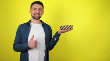 Beyaz tişörtlü genç bir adam avucunda bir hediye tutuyor, gülümsüyor ve sarı arka plan gibi görünüyor. Tatil konsepti: Yeni yıl, Noel, Aziz Nicholas Günü, Sevgililer Günü, doğum günü. 4k görüntü