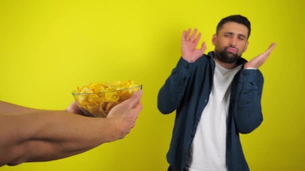一个身穿蓝色衬衫和白色T恤的年轻人拒绝了一个装有薯片和垃圾食品的玻璃器皿的求婚 健康饮食的概念 黄色背景 有复制空间 4K慢镜头 — 图库视频影像
