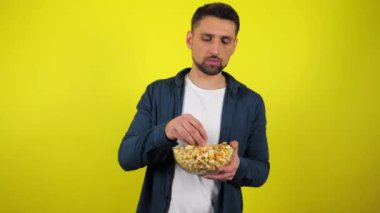 Mavi tişörtlü ve beyaz tişörtlü genç adam şeffaf cam tabaktan lezzetli patlamış mısır yemiyor ve bundan hoşlanmıyor. Bozuk bakkaliye. Abur cubur konsepti. Kopyalama alanı olan sarı arkaplan. 4k yavaş çekim