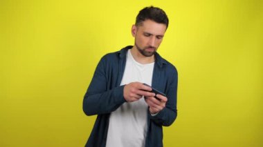 Mavi tişörtlü ve beyaz tişörtlü genç bir adam akıllı telefonda oyun oynuyor ve kaybediyor, üzülüyor. Kopyalama alanı olan sarı arkaplan. 4k yavaş çekim görüntüsü