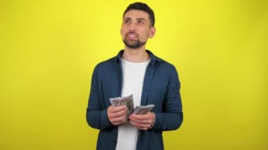 Mavi tişörtlü ve beyaz tişörtlü genç bir adam bir paket para sayıyor ve satın almayı planlıyor. Kopyalama alanı olan sarı arkaplan. 4k yavaş çekim görüntüsü