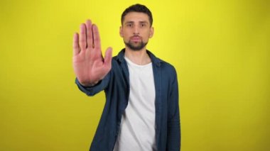 Mavi tişörtlü ve beyaz tişörtlü genç adam kameraya bakar, elini uzatır ve dur der. Kıpırdamayın. Kopyalama alanı olan sarı arkaplan. Hareketi durdurun. 4k yavaş çekim görüntüsü.