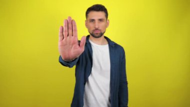 Mavi tişörtlü ve beyaz tişörtlü genç adam kameraya bakar, elini uzatır ve dur der. Kıpırdamayın. Kopyalama alanı olan sarı arkaplan. Hareketi durdurun. 4k yavaş çekim görüntüsü.