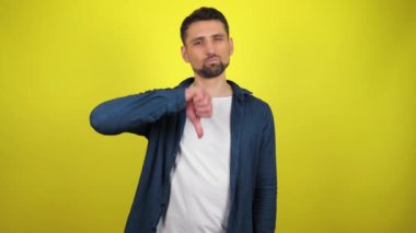 Mavi tişörtlü ve beyaz tişörtlü genç bir adam ciddi olarak kameraya bakıyor ve elle nefret gösteriyor. Kopyalama alanı olan sarı arkaplan. 4k yavaş çekim görüntüsü.