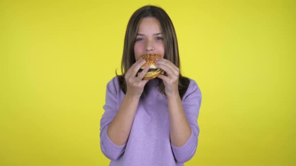 ピンクのプルオーバーの10代の女の子がハンバーガーを食べてカメラを見ている コピースペース付きの黄色の背景 ジャンクフード健康的な食事の概念 10代の感情4Kスローモーション映像 — ストック動画