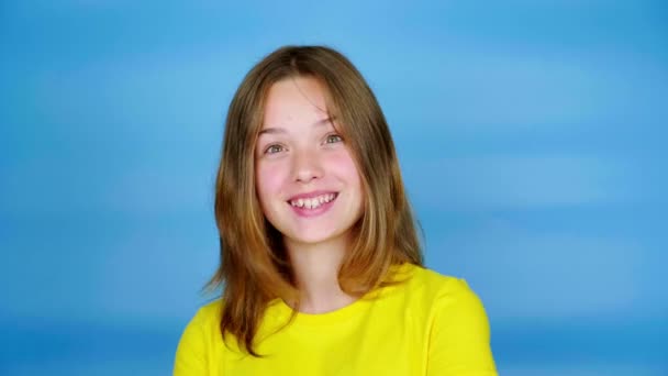 穿着黄色T恤的快乐少女看着摄像机 点点头说是的 蓝色背景 有复制空间 青少年的情绪 4K镜头 — 图库视频影像
