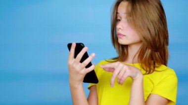 Sarı tişörtlü genç kız akıllı telefondan kayıyor, şaşırıyor ve vay canına diyor. Kopyalama alanı olan mavi arkaplan. Gençlik duyguları. 4k görüntü