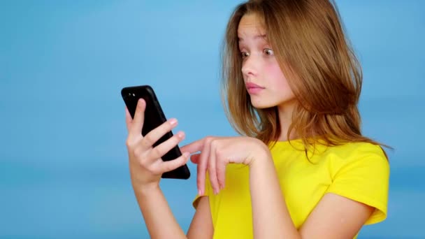 黄色のTシャツの10代の少女は スマートフォン上でスワイプされ 驚きとすごいと言う コピースペース付きの青の背景 10代の感情4K映像 — ストック動画