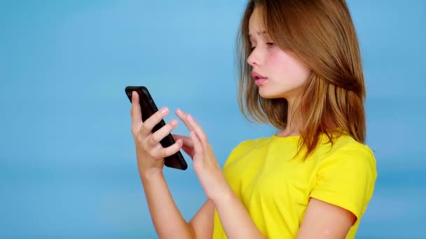 黄色のTシャツの10代の女の子は スマートフォン スワイプタッチスクリーンで使用されます コピースペース付きの青の背景 10代の感情4K映像 — ストック動画