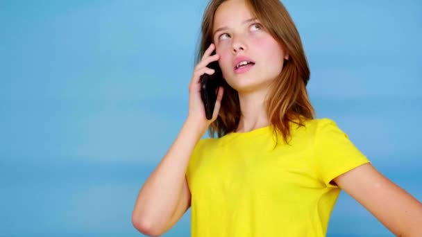 黄色のTシャツを着た10代の少女がスマートフォンを呼び出し 話し始める コピースペース付きの青の背景 10代の感情4K映像 — ストック動画