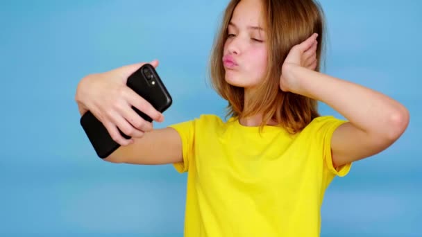 黄色のTシャツを着た幸せな10代の女の子は スマートフォンや笑顔で自撮りやビデオを作ります コピースペース付きの青の背景 10代の感情4K映像 — ストック動画